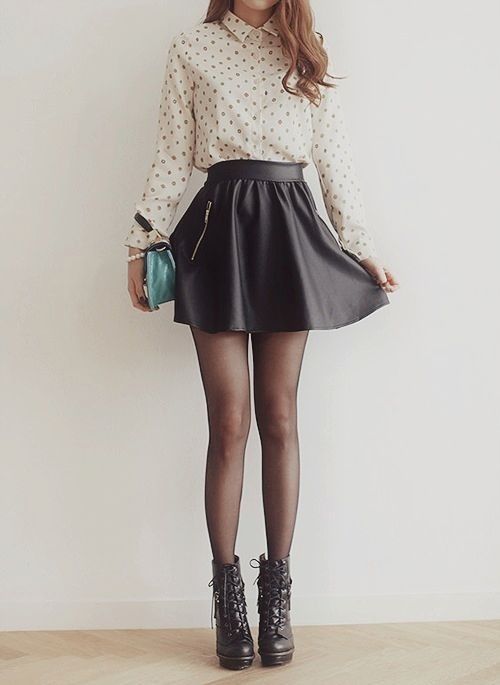 a woman is wearing a white polka dot shirt, a black skirt, black tights, and black lace up boots. she has a small turquoise bag and is looking at the camera.  she is standing in front of a white wall.
