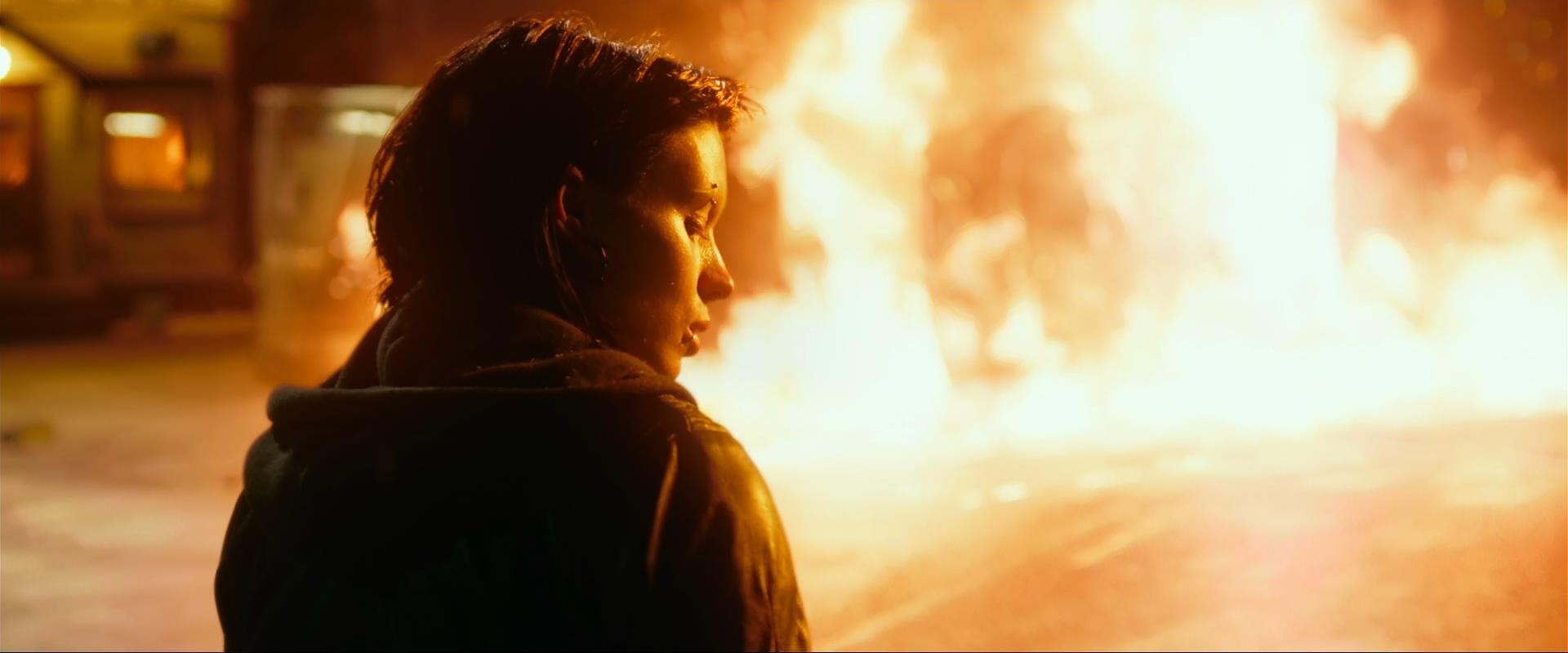 a young woman stands in the foreground of the image, silhouetted against a bright, outoffocus background of fire. she is looking away from the viewer, with a somber expression on her face. the fire appears to be burning in the street behind her, and the scene is bathed in an orange glow. the image conveys a sense of danger, sadness, and despair.