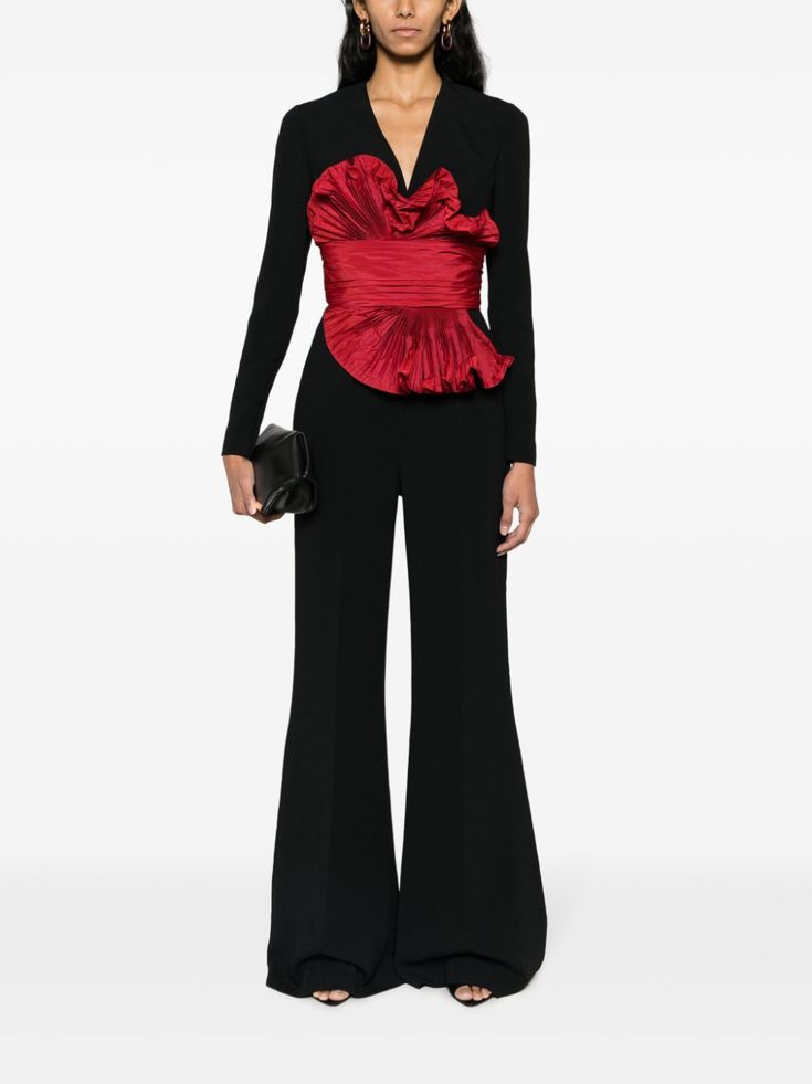 a woman model is wearing a black jumpsuit with a red ruffle bodice. she is standing in a studio setting with a white background. she is posing with her hands on her hips. she is wearing a black clutch.