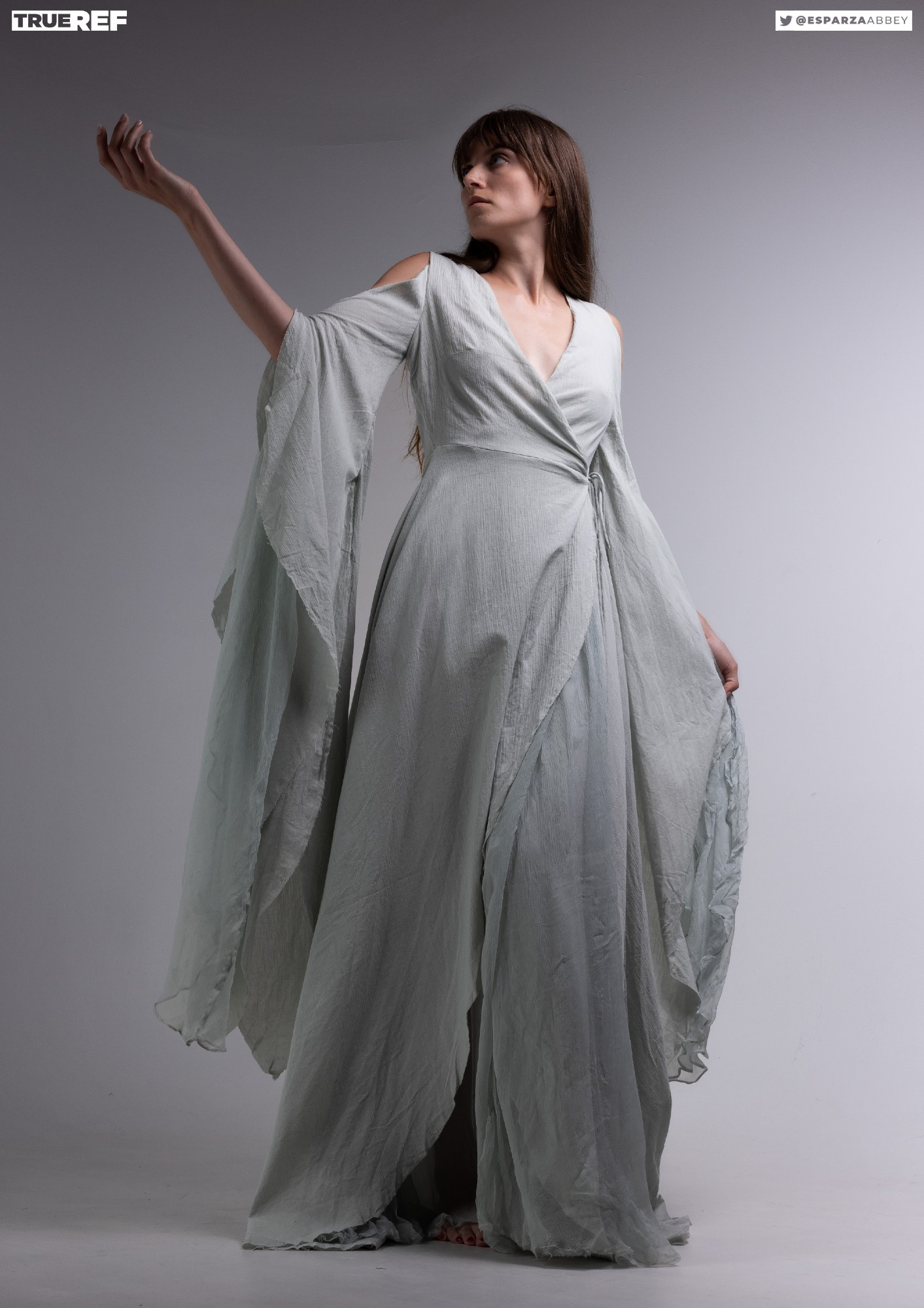 a fulllength studio shot depicts a woman model gracefully posing in a flowing, light sage green wrap dress. the dress features long, wide sleeves that drape dramatically around her.  the style is ethereal and romantic, with soft, delicate fabric. the woman's pose is elegant and suggestive of movement. the background is a simple, light grey, allowing the dress and model to be the focus. the watermark "@esparzaabbey" and "true ref" are visible.