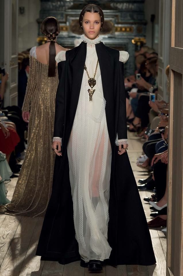 a runway scene captures a female model walking towards the camera, wearing a white lace dress that is partially sheer, paired with a long black coat. she has a ruffled collar and a large gold necklace. in the background, another model in a gold dress walks away. spectators line the sides of the runway, taking photos.