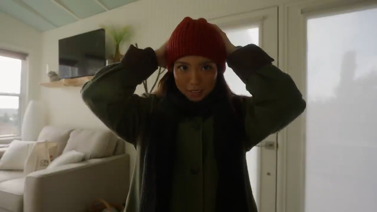 a young woman stands in a living room, putting on a red beanie. she wears a green jacket and a black scarf. the room is bright with natural light coming from a window on the left side. the living room is furnished with a beige sofa, a wooden coffee table, and a large flatscreen tv mounted on the wall. the overall ambiance is warm and inviting.