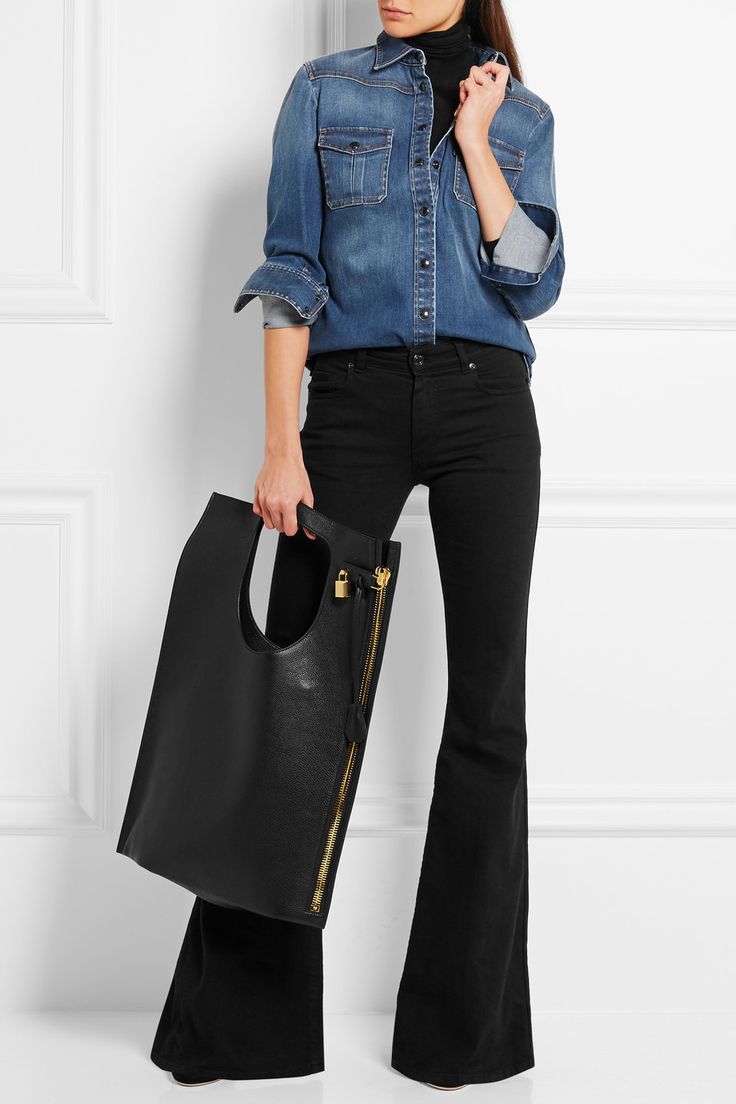 a woman is standing in front of a white wall, wearing a denim shirt over a black turtleneck, black flared pants, and holding a large black handbag with gold zipper detailing.