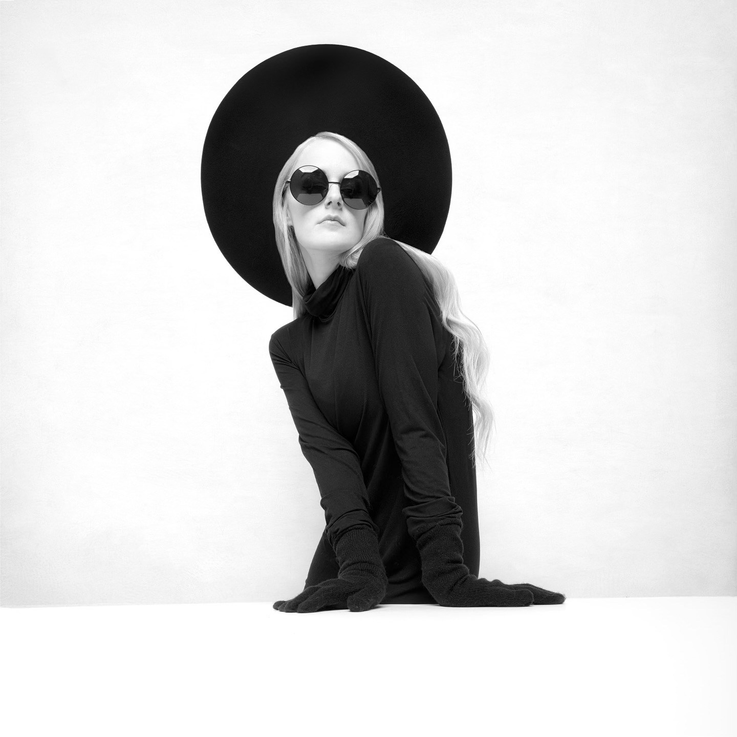 a woman in a black turtleneck sweater and sunglasses, wearing a large black hat, poses with a serious expression. the background is a white wall, creating a striking contrast with her black outfit. the image is in black and white, emphasizing the minimalist fashion statement and creating a sense of mystery. the woman's pose is elegant and confident, suggesting a strong and independent personality.  the image is a compelling example of fashion photography, showcasing a modern and timeless aesthetic.