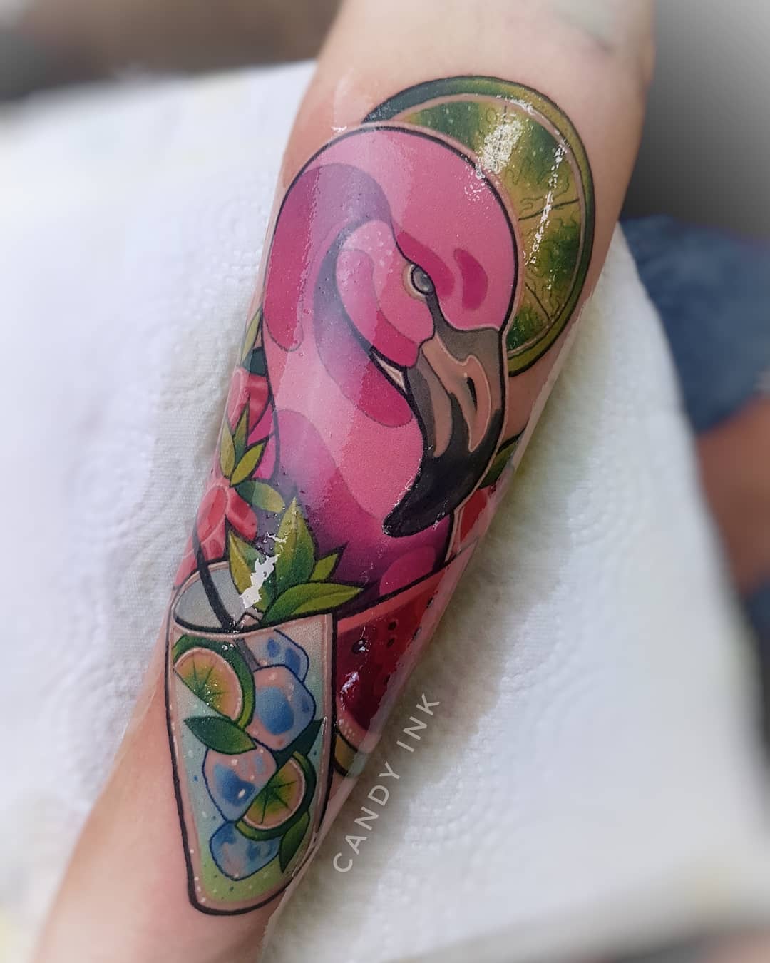 a person's arm with a colorful, detailed tattoo of a flamingo, cocktail, watermelon, and lime slices. the tattoo has a cartoonish style, with bold lines and vibrant colors.  the tattoo is on the inner forearm, extending towards the wrist.