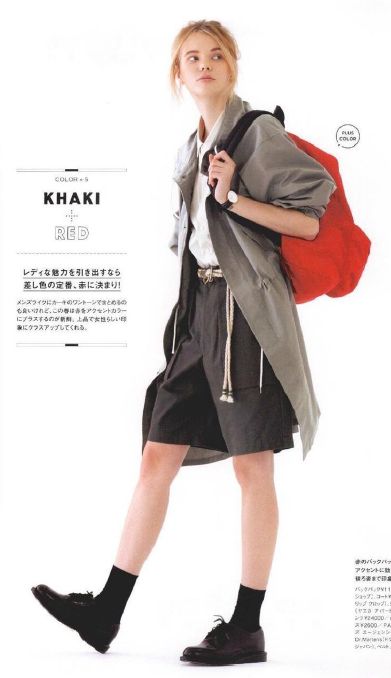 a young woman is wearing a khaki jacket and red bag,  she is wearing black shorts with a  black belt and black  shoes.  she is standing on a white background.  the image is a fashion editorial.
