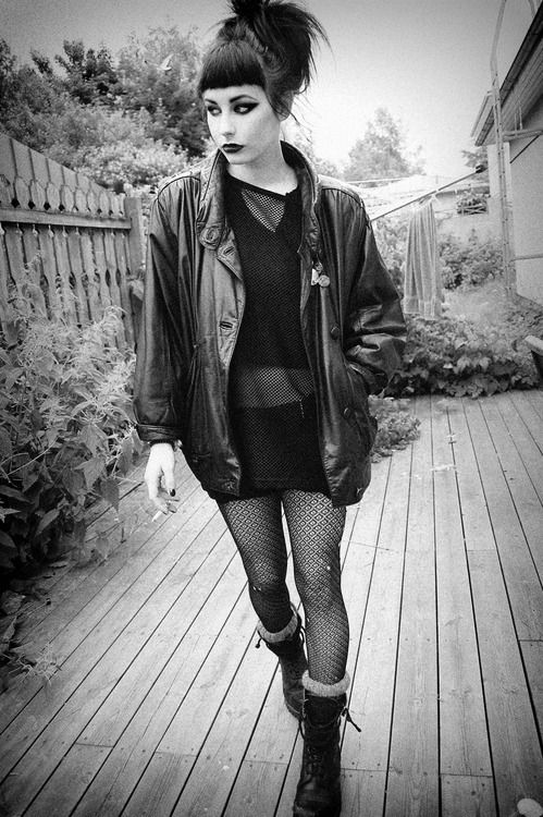 a black and white full shot of a young woman with short, dark hair and bangs, dressed in punk/goth style clothing. she's wearing a black leather jacket, fishnet stockings, and combat boots. she's walking on a wooden deck in a backyard, with a fence visible in the background. she seems to be holding or just finished smoking a cigarette. the overall mood is dark and alternative.