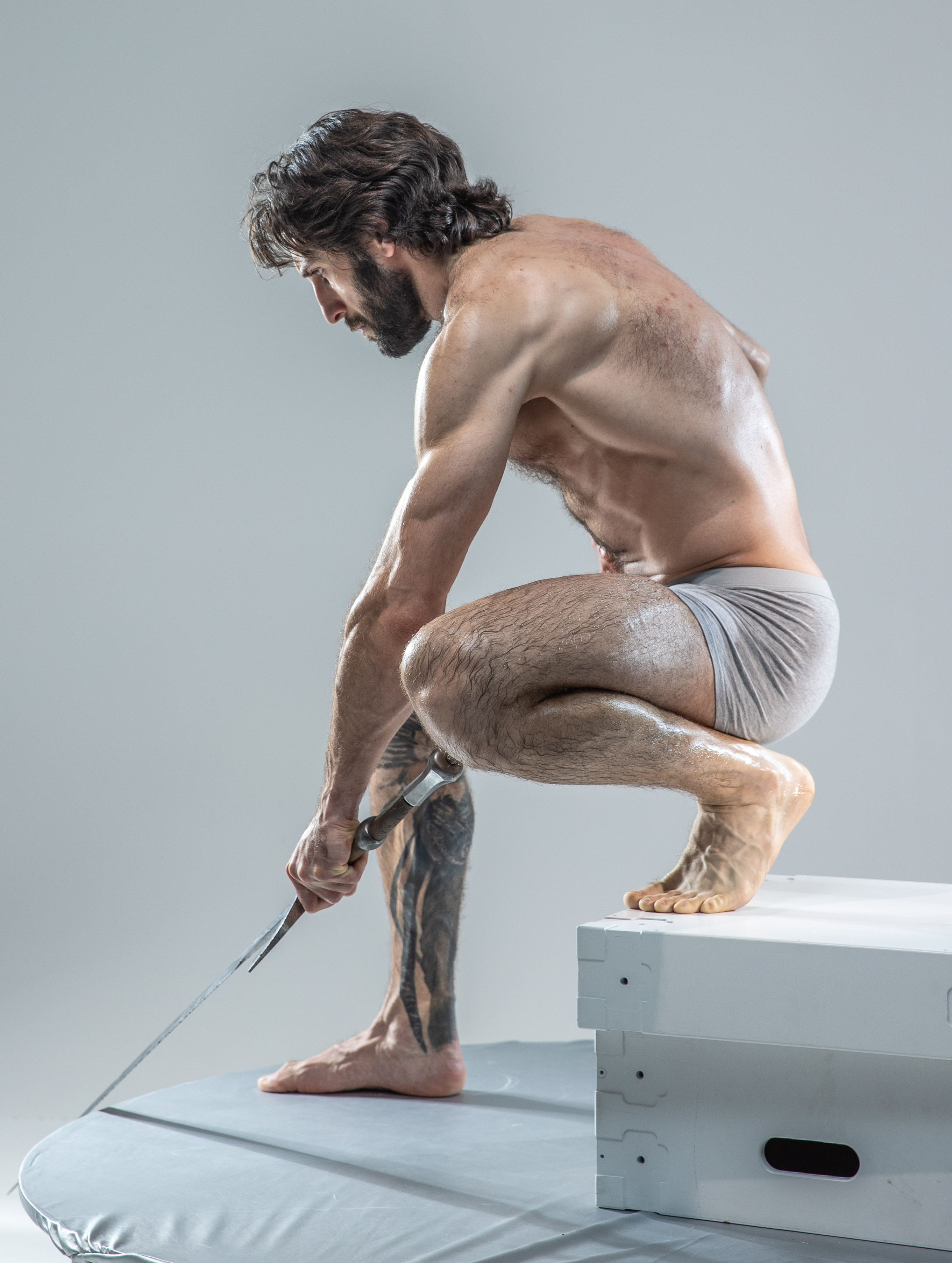 a muscular, bearded man in grey underwear is posed in a studio setting. he is crouched on a white platform, gripping a hammer in his right hand and a nail in his left. his body is angled to the side, showcasing his physique and the tension in his muscles. the lighting is soft and even, highlighting the contours of his body. the background is a plain, light grey.