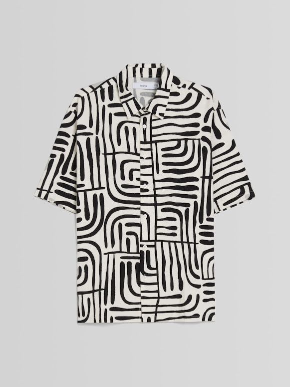 a shortsleeved buttondown shirt with a black and white geometric pattern is displayed. the pattern consists of thick, curved and straight lines that create an abstract design. the shirt is white with black lines. a tag is visible on the collar.