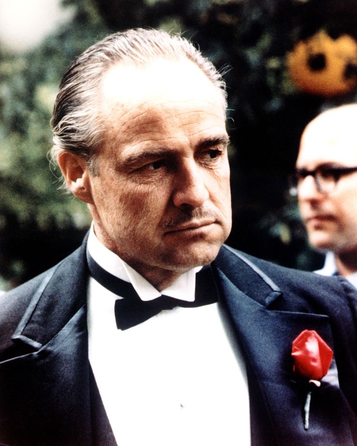 a portrait of marlon brando as don corleone in the godfather. he is wearing a black suit with a white shirt and bow tie, and a red rose on his lapel. another man with glasses is partially visible behind him.
