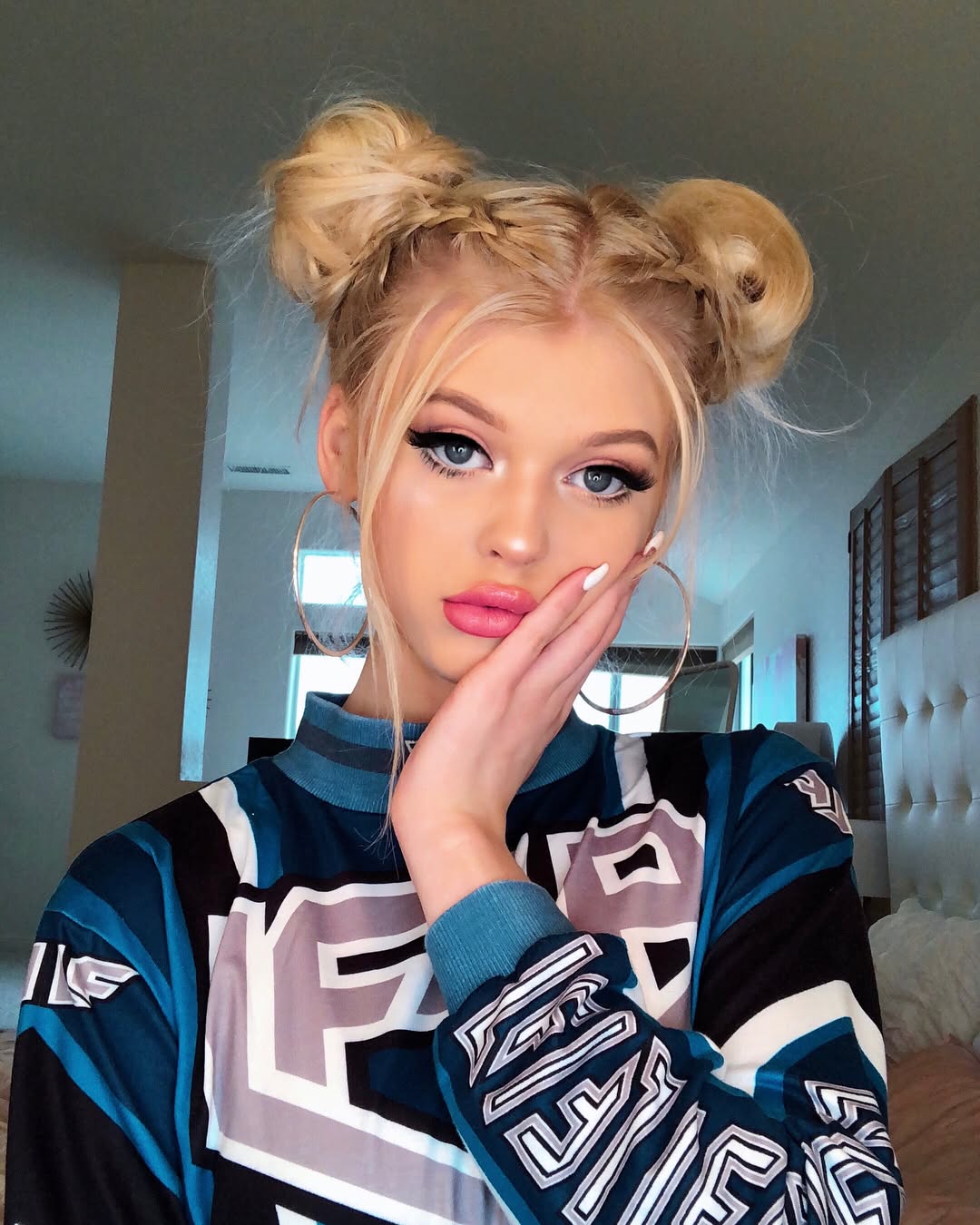 a blonde woman with her hair styled in space buns with braids poses for a selfie. she is wearing hoop earrings, makeup, and a longsleeved sweater. her hand is on her face and her nails are painted white. the photo was taken indoors.