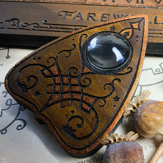 a closeup shot of a handcrafted ouija board planchette. the planchette is made of wood and is painted in shades of brown, black, and copper. it has a circular window in the center and is decorated with intricate carvings. the planchette is resting on a piece of paper with a vintage map background.