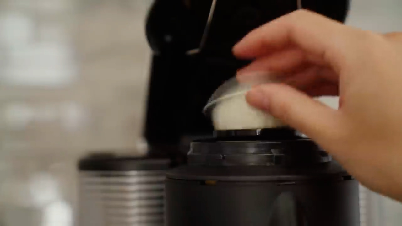 a closeup shot of a person's hand inserting a singleserve coffee pod into a black coffee maker. the coffee pod is white with a silver ring around the top. the coffee maker is made of black plastic and has a silver water tank on the left side. the background is a blurry white tiled wall.
