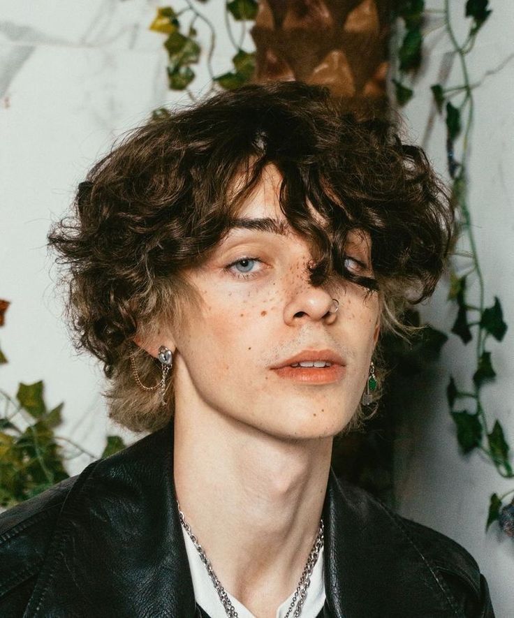 a closeup portrait of a young man with curly brown hair and freckles. he has a nose piercing and wears multiple earrings and necklaces. he is wearing a black leather jacket.  the background is blurred and features a white wall with green ivy.  the lighting is soft and natural, highlighting his features.
