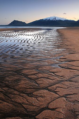 a stunning image of a beach landscape at sunset.  the sand is smooth and even, with a pattern of waves and lines, and the water is calm and still, with a beautiful sunset sky.  the mountains rise up in the background, creating a sense of depth and awe.  the beauty of nature is evident in this picture.