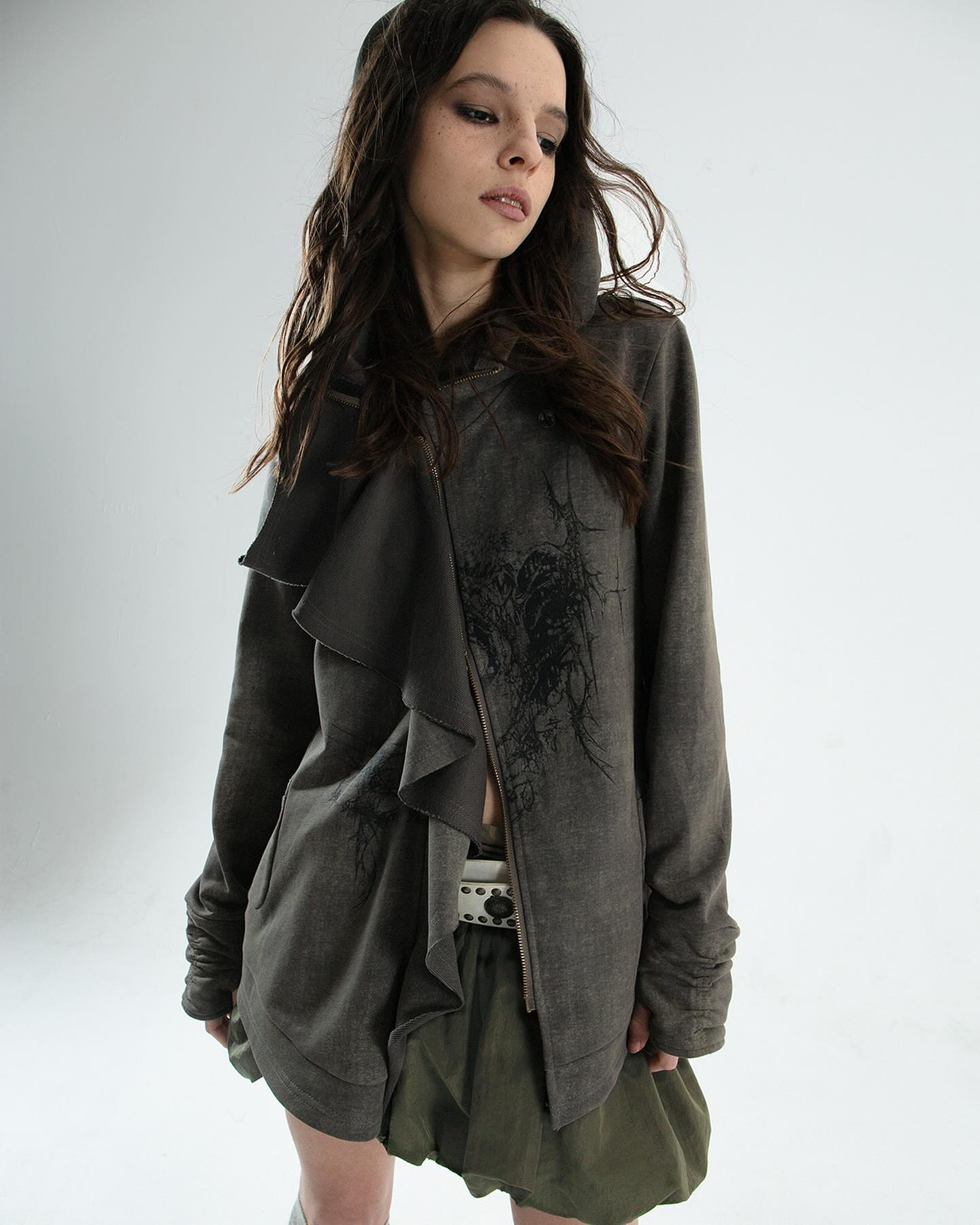 a young woman with long brown hair models a grey hooded jacket with a ruffled front detail and an olive green skirt.  the jacket has a distressed or grungelike appearance with what appears to be a dark ink design. the photo is taken against a plain white background. the model appears serious and somewhat melancholic. the overall style is casual but edgy.