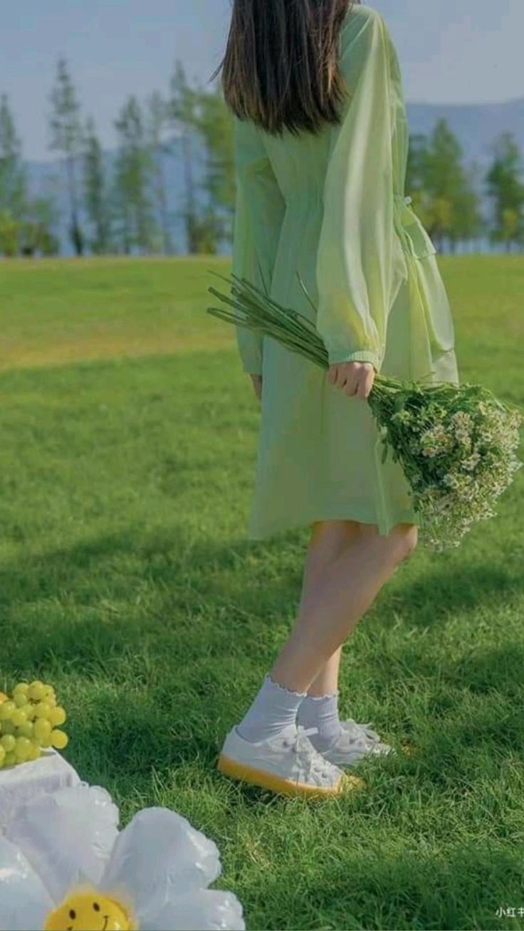 a woman in a flowing green dress strolls through a vibrant green field under a bright blue sky. she holds a bouquet of white wildflowers and wears white sneakers. the perspective is from behind, capturing her leisurely movement.  a picnic setup with grapes and an inflatable smiley face decoration is visible in the foreground, suggesting a joyful and carefree atmosphere.
