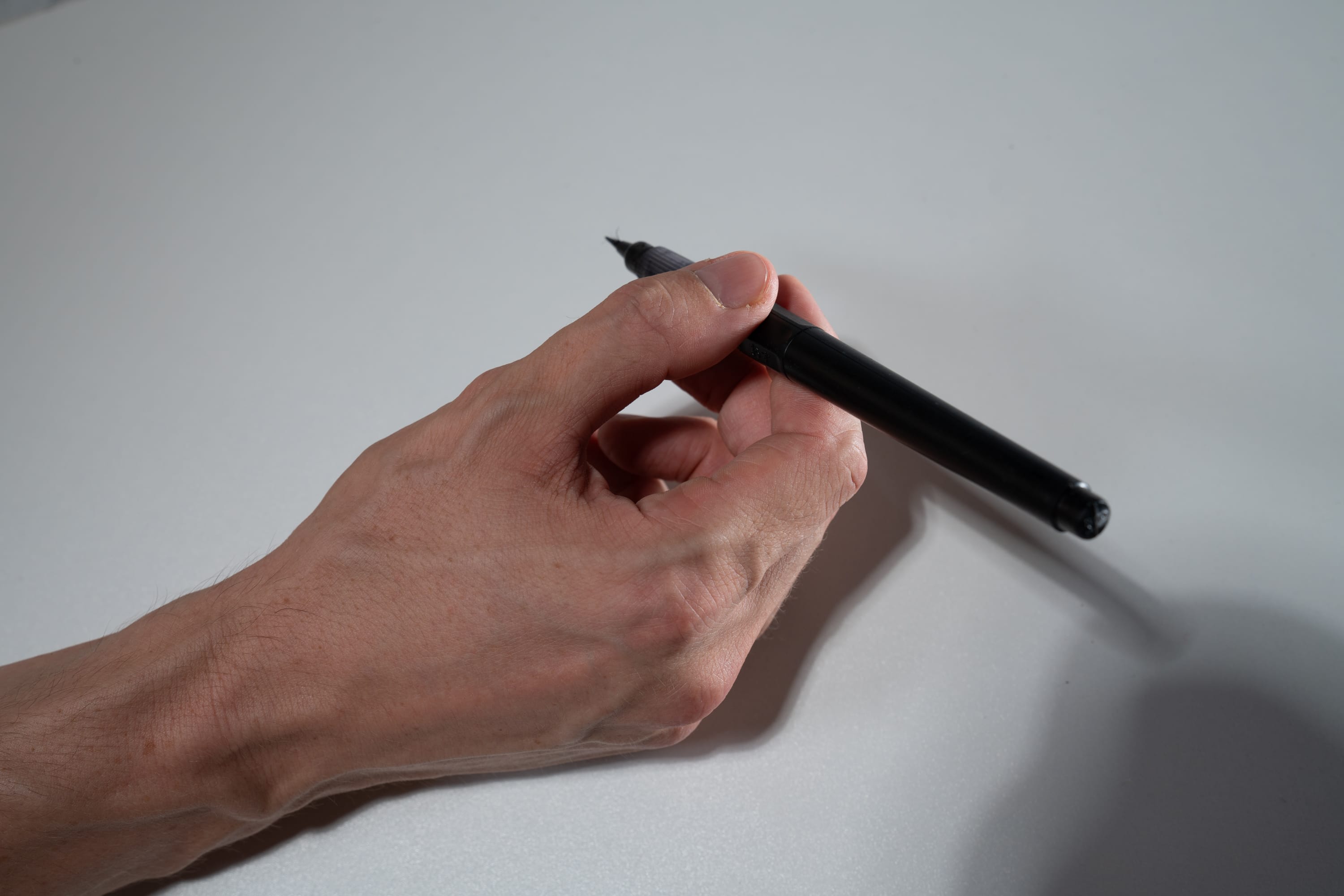 a hand is holding a black pen against a white background. the hand is positioned as if it is about to draw or write something. the lighting is natural, creating a soft shadow on the background.