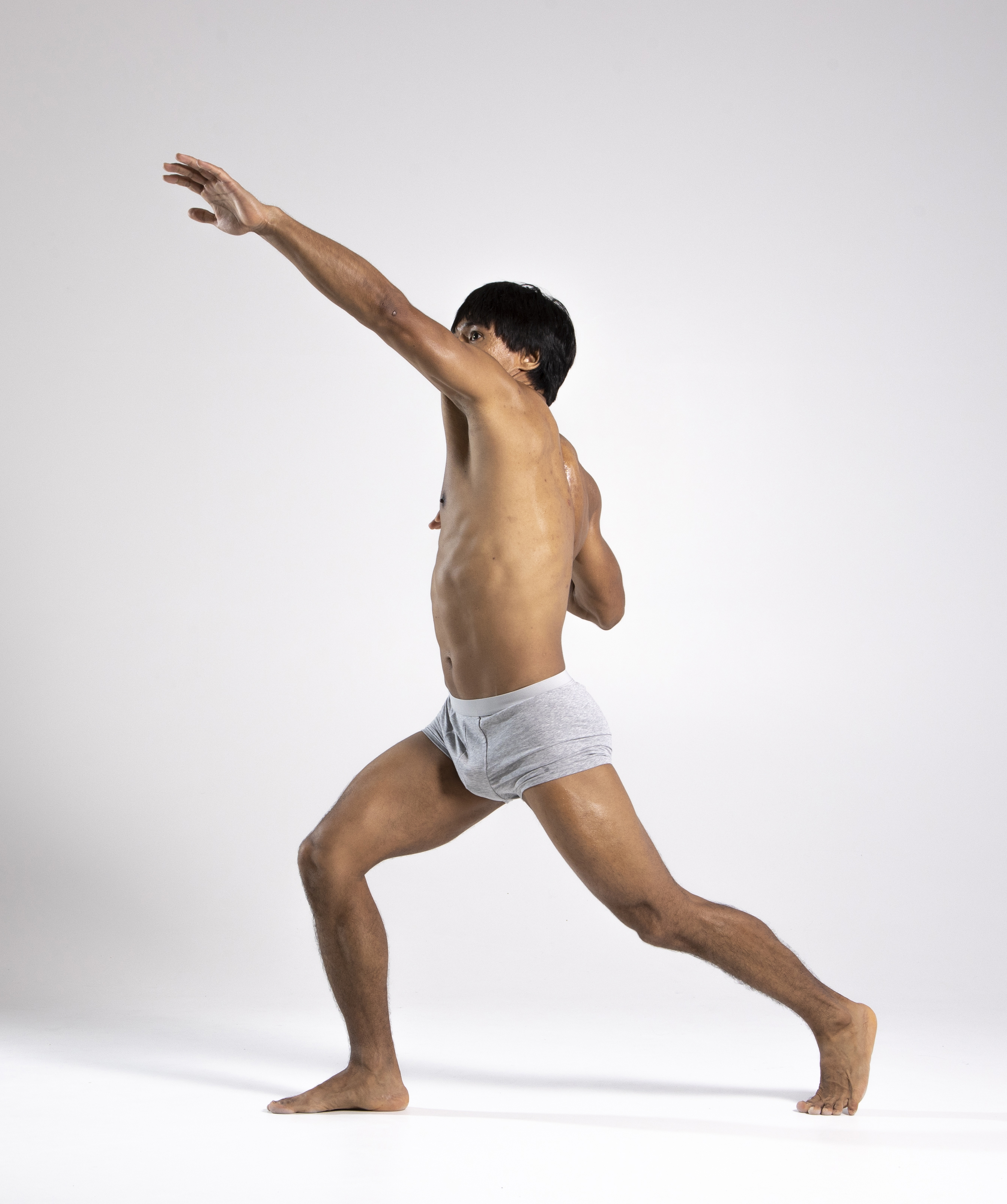 a shirtless man in gray underwear is captured in a full shot against a white studio background. he is in motion, either stretching or exercising, with his arm extended upward. his posture is athletic and dynamic.