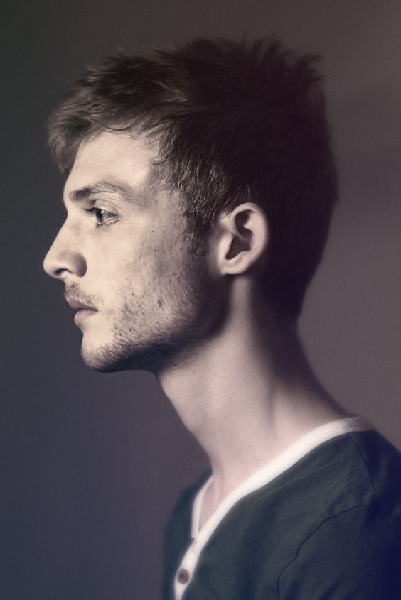 a profile portrait of a young man with short brown hair and light stubble. he is wearing a green button down shirt with a white collar. the background is a blurred gray.