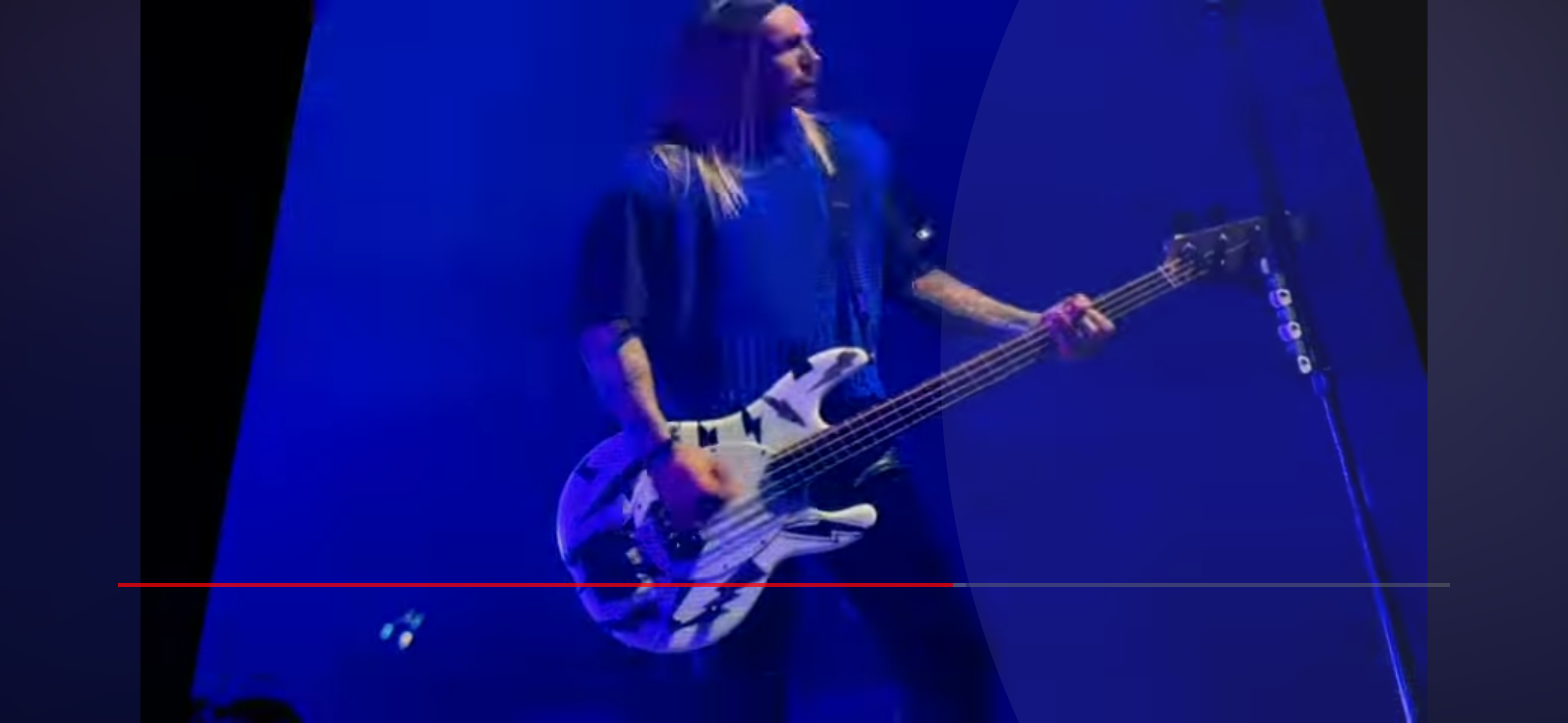 a musician is playing a bass guitar on stage during a concert.  the performer is wearing a black shirt with long hair. the stage is lit in blue light and a microphone stand is visible to the right. the focus is on the guitar and the musician's hand as they play.
