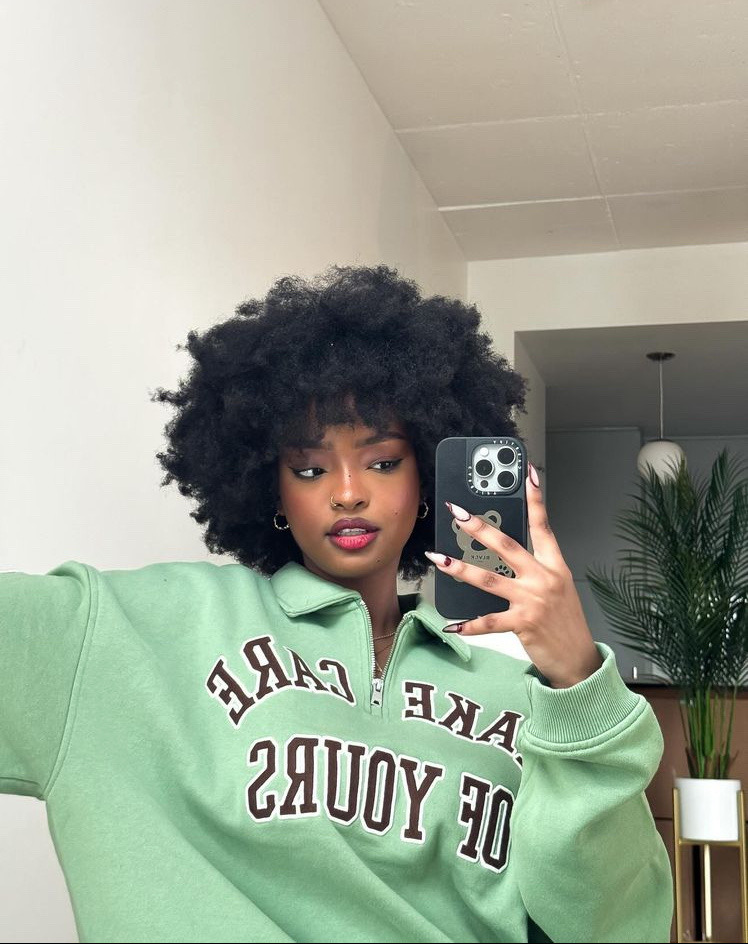 a young black woman with beautiful afro curls is taking a selfie while wearing a mint green sweatshirt that says "take care of yours" on the front.  she is looking at the camera and smiling.