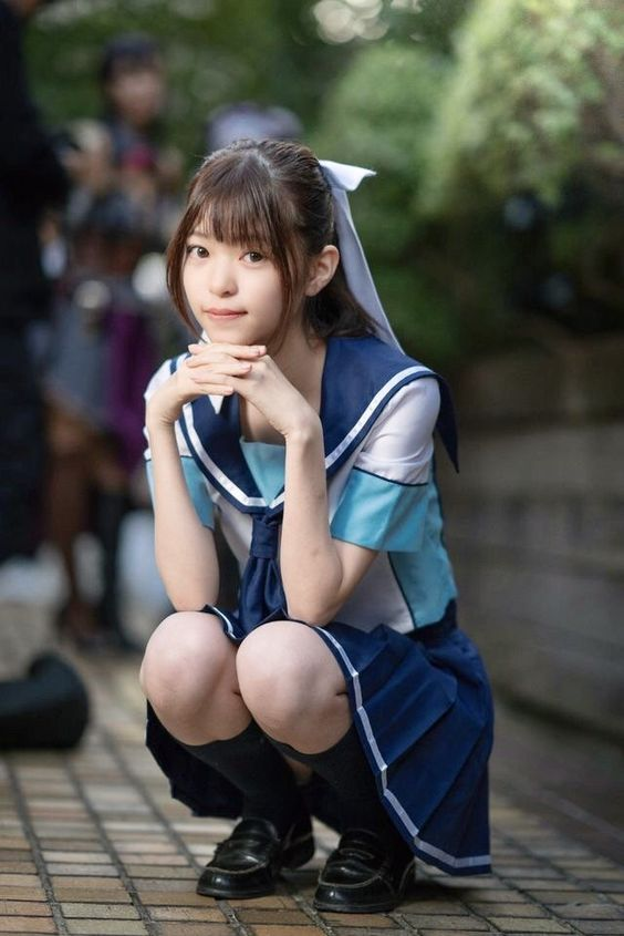 a young japanese woman dressed in a blue and white sailorstyle uniform squats on a brick sidewalk while looking directly at the camera. she has a serene expression on her face and her hands are clasped beneath her chin. the background is slightly blurred and shows other people and greenery, suggesting an outdoor setting. the lighting appears to be natural and slightly overcast.