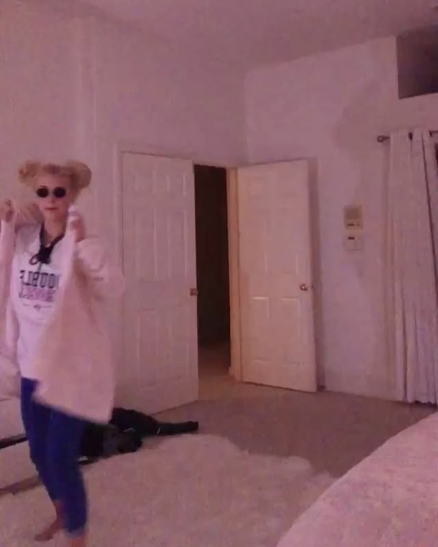 an indoor shot of a person standing in a bedroom. the person has blonde hair in space buns and is wearing sunglasses, a white tshirt with "doubt" printed on it, a pink jacket, and blue pants. they are standing on a shag rug, facing forward, with a doorway visible behind them. the doorway leads to another room that's dark inside. to the right of the doorway is a white door, closed. a beige curtain hangs on the right, and part of a bed is visible in the bottom right.