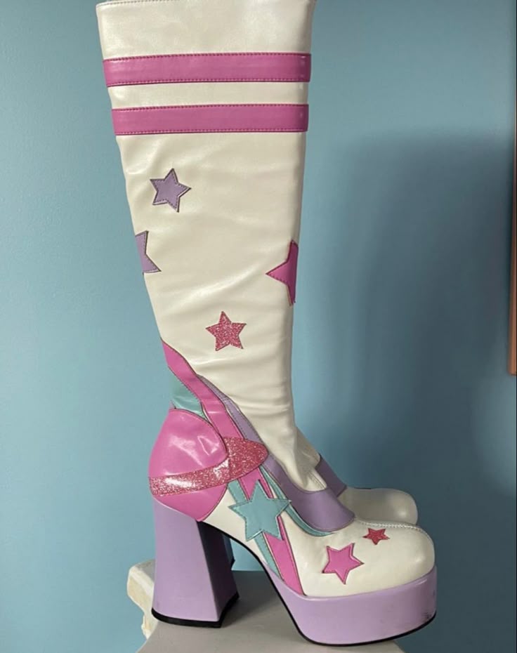 a single white kneehigh platform boot with pink, purple, and blue accents is displayed against a light blue wall. the boot has two horizontal pink stripes near the top and various stars in different sizes and colors scattered on the shaft. the heel and platform are purple, and there are additional pink and blue decorative elements around the ankle and foot area.