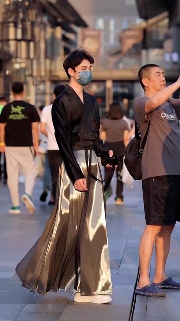 a person wearing a traditional black and silver hanfu stands on a busy street with other people walking in the background. he wears a light blue surgical mask, and another man next to him holds up a phone to take a picture.