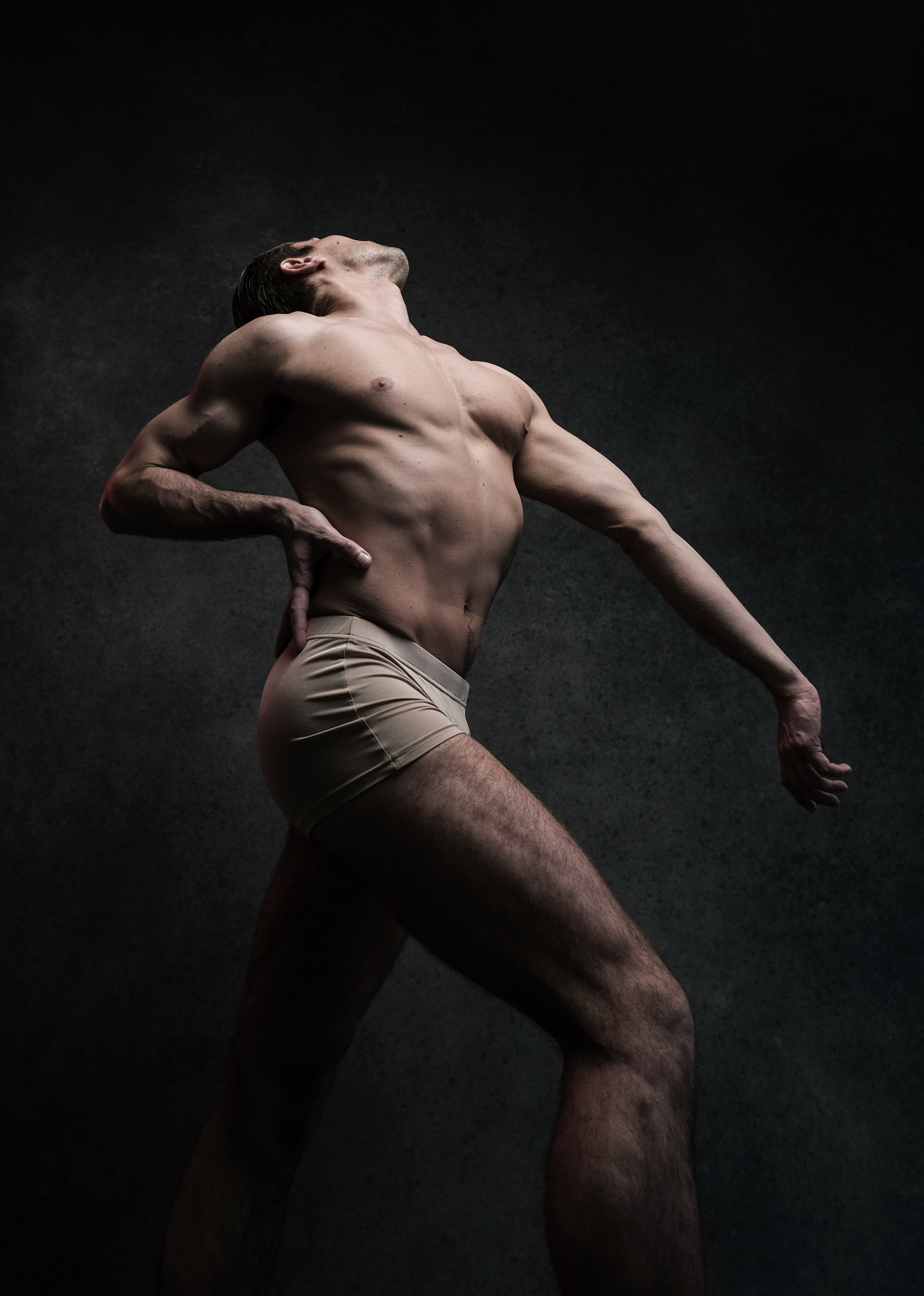the image showcases a male dancer in a dynamic pose against a dark grey background. the lighting emphasizes his muscular physique and the artistic expression of the pose. his body is contorted in a way that suggests movement and power, creating a sense of energy and grace. the dancer's gaze is directed upwards, adding a layer of emotional intensity to the image.