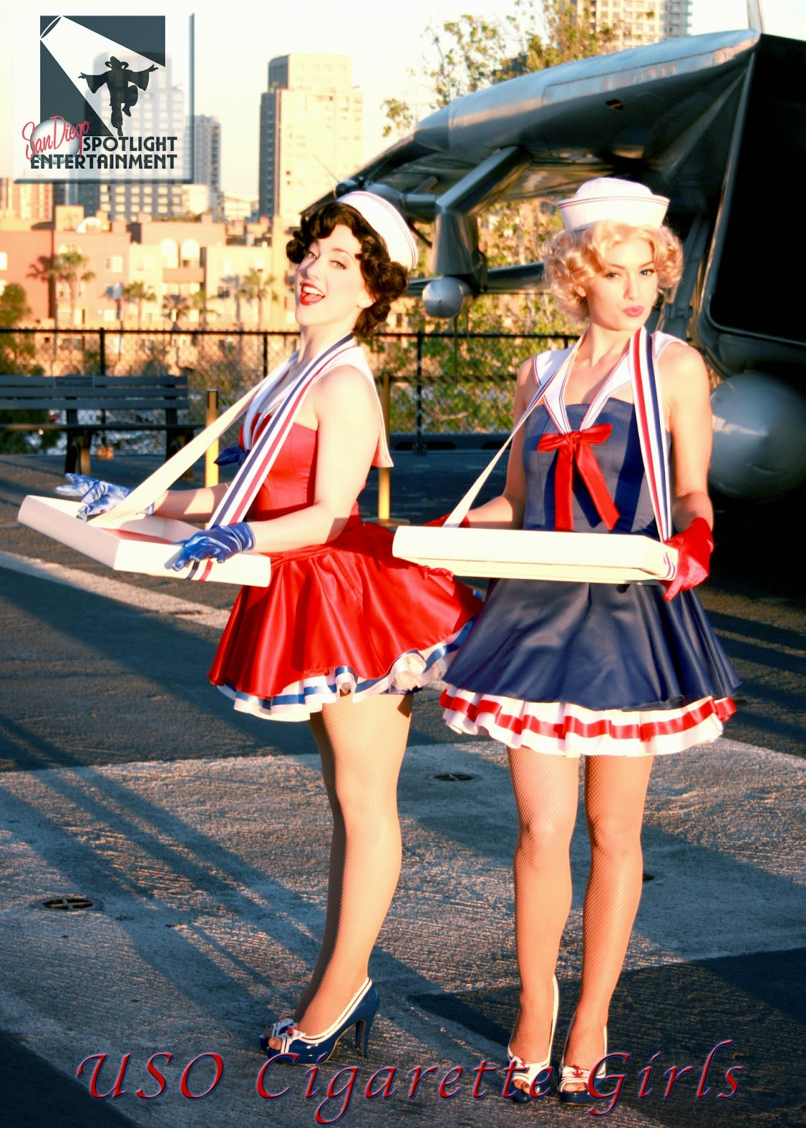 the image depicts two women dressed in vintagestyle sailor girl costumes reminiscent of uso cigarette girls from the mid20th century.  they are wearing red, white, and blue outfits with sailor hats and are carrying trays. the setting appears to be an aircraft carrier or airfield, enhancing the historical context.  a logo for "san diego spotlight entertainment" is visible in the top left corner. the overall aesthetic is that of a nostalgic pinup or historical recreation, perhaps for a performance or photoshoot.  the bright sunlight and shadows cast on the tarmac contribute to the overall mood and setting.