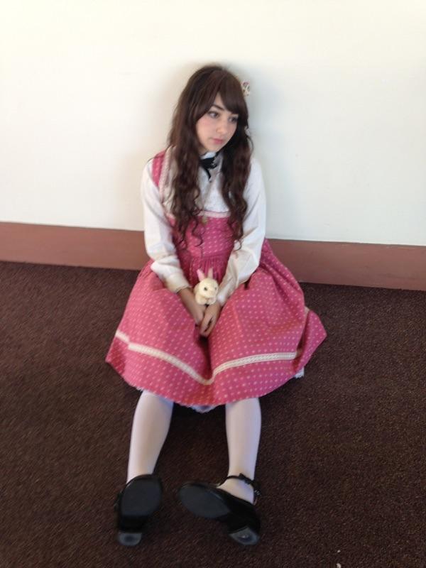 a young girl sits on a dark brown carpet against a light beige wall. she is wearing a pink polka dot dress over a white blouse, white socks, and black shoes. she is holding a small white rabbit doll in her lap. her long brown hair is partially covering her shoulders. she is looking off to the side with a slightly wistful expression. the overall style is reminiscent of lolita fashion.