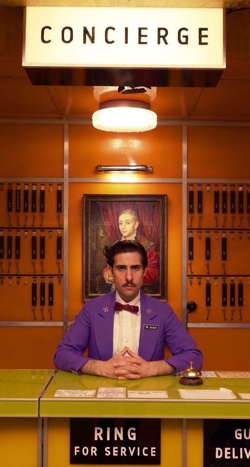 a concierge stands behind a front desk in a hotel lobby. the concierge has a mustache and is wearing a purple jacket and bow tie. the desk is made of wood and has a sign that reads "ring for service." behind the concierge is a vintage painting of a man in a suit. there are keys hanging on the wall behind the concierge. the room is decorated in orange, yellow, and green.  the concierge is waiting for guests.
