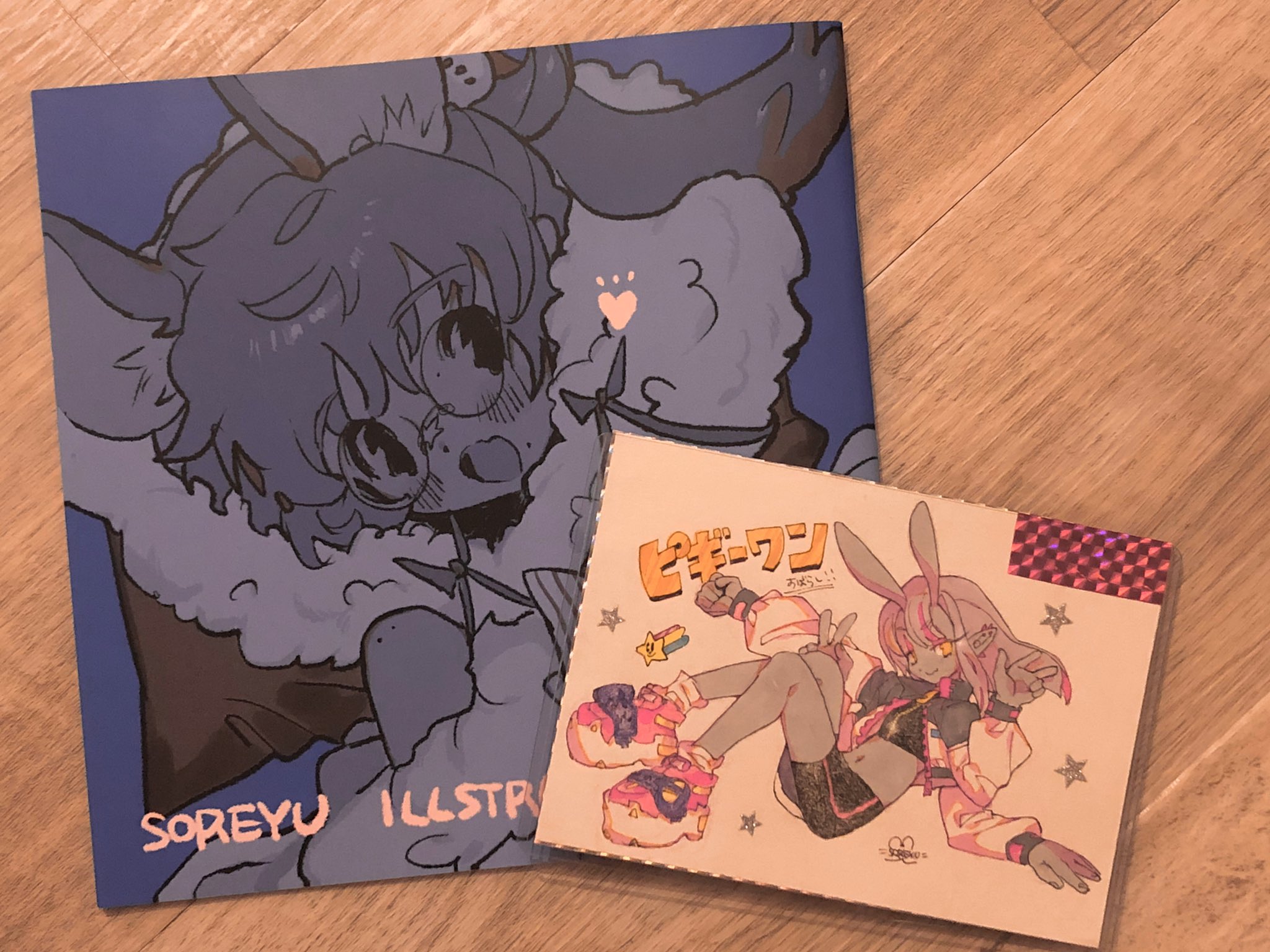 two squarebound illustration zines lay on a light brown wood floor. the larger zine is mostly blue and features a character with deer ears and antlers. the smaller zine is primarily white and features a character with rabbit ears. both characters have colorful accents.