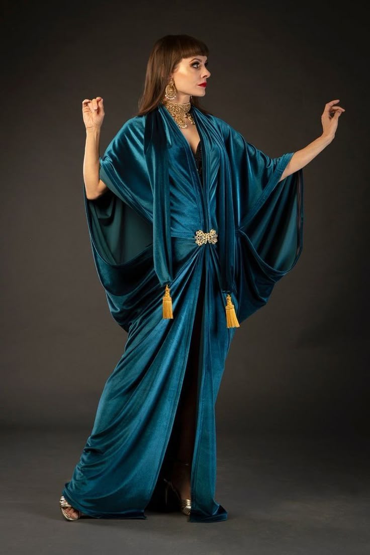 a fulllength studio shot features a woman in a striking teal velvet gown. the dress has a deep vneckline, cinched at the waist with a decorative gold brooch and gold tassels. the gown drapes elegantly, with wide sleeves that add a touch of drama. the woman accessorizes with gold jewelry including large earrings and a statement choker. she's standing against a dark gray backdrop, posed with one arm raised.