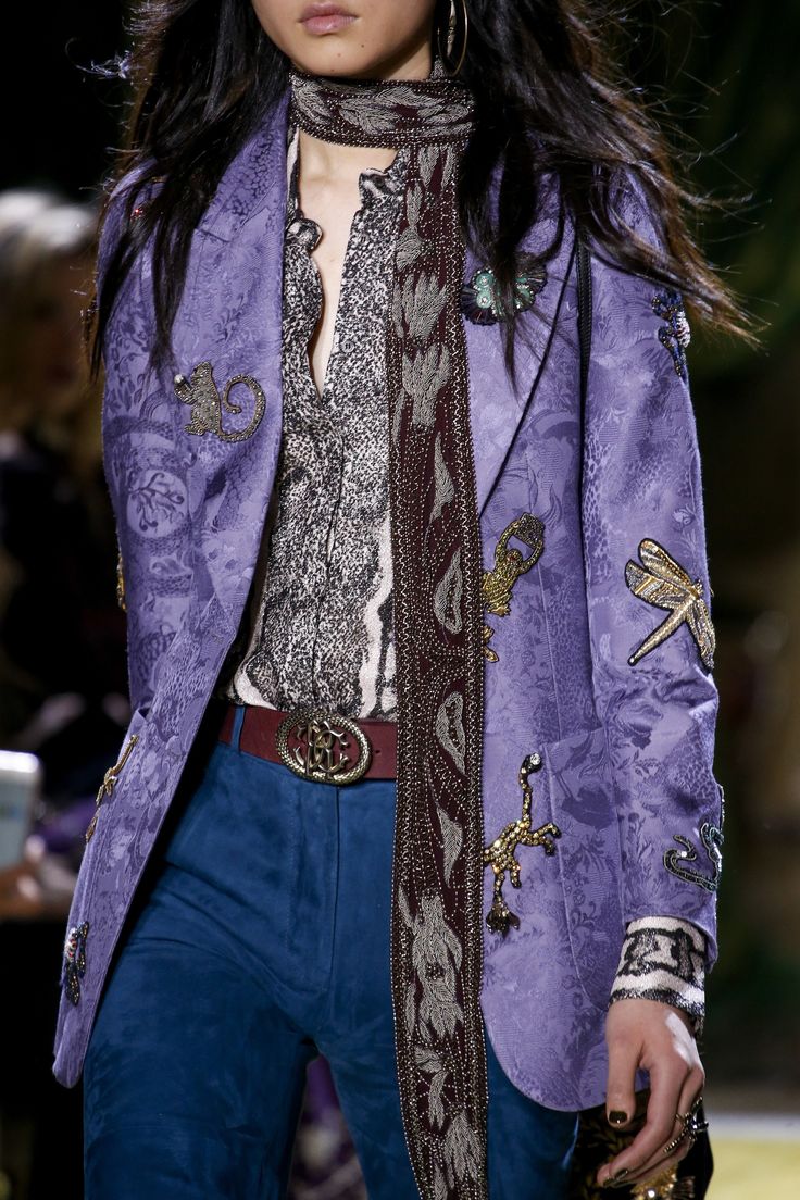 closeup shot of a female model on a fashion runway, showcasing a stylish purple embroidered jacket with various embellishments like dragonflies and other creatures. the jacket is worn over a gray patterned shirt and paired with dark blue suede pants and a burgundy beaded scarf. a belt with a noticeable buckle is also visible. the model's dark hair is styled loosely around her shoulders, and subtle makeup enhances her features. the background is blurred but suggests a runway setting. the image focuses on the intricate detailing and rich textures of the clothing.