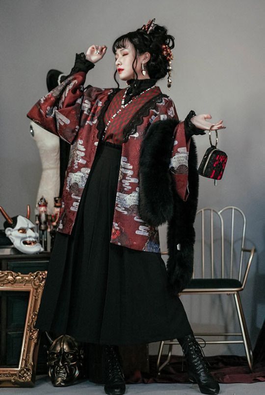 a young woman is posing in a studio. she is wearing a dark red and black kimonostyle jacket over a black hakama skirt. the jacket is decorated with a traditional japanese pattern. she is also wearing black laceup boots and has her hair styled in a traditional updo. the background is dark and moody, with a chair and some traditional japanese masks visible. she has a black fur stole draped over her shoulders and is holding a small black bag.  the overall style is a blend of traditional japanese fashion and dark, gothic lolita elements.