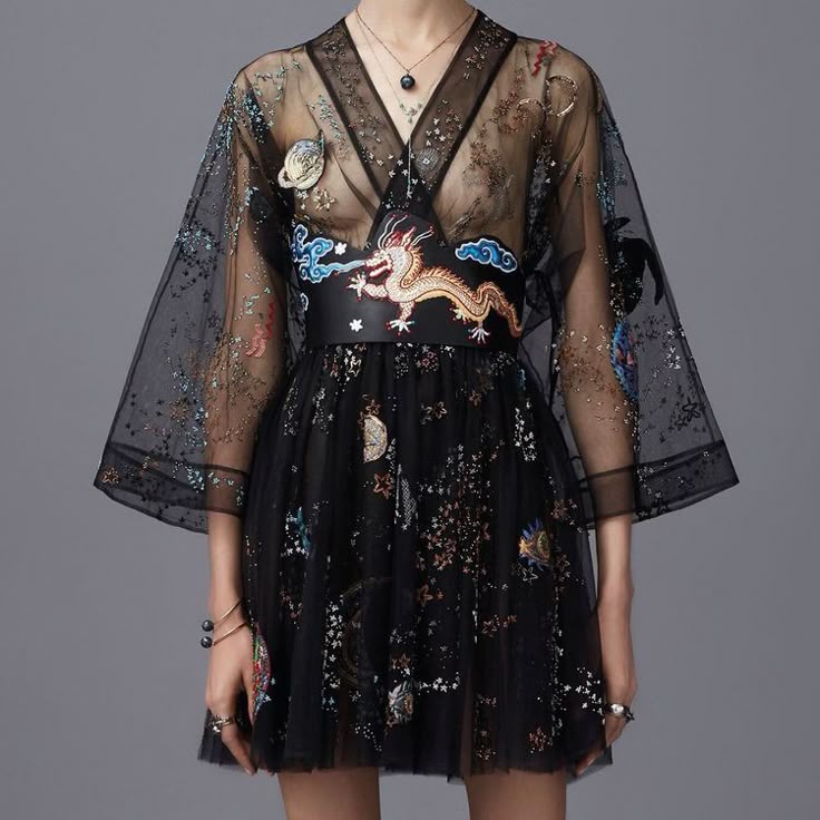 a woman is wearing a black, sheer, vneck dress with kimonostyle sleeves and a black belt embroidered with a dragon. the dress features embroidery with spacethemed designs. she is also wearing a necklace and jewelry. the background is a solid gray color.