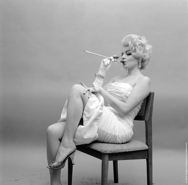 a black and white photo of a glamorous woman in a white dress sitting on a chair. she is holding a cigarette holder and appears to be smoking. she is wearing high heels, gloves, and jewelry. the photo has a vintage feel, likely from the 1960s. the background is a simple, lightcolored studio backdrop. the woman is styled and posed in an elegant manner.