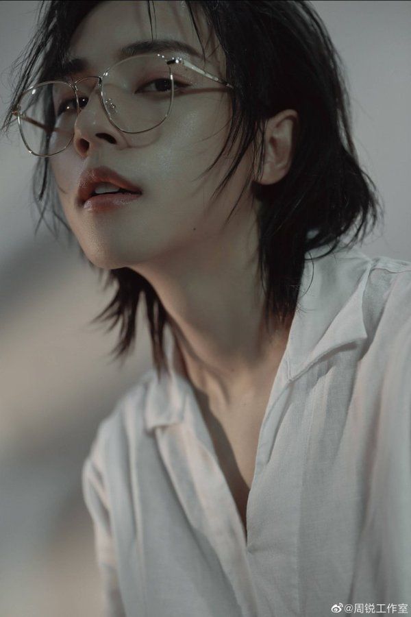 the image shows a closeup portrait of a young woman with short, dark hair styled in an androgynous fashion. she is wearing clear, thinframed glasses and a slightly unbuttoned white collared shirt. the lighting is soft and diffused, creating a gentle highlight on her skin and accentuating her delicate features. her gaze is directed slightly upwards, engaging the viewer with a captivating intensity. the overall aesthetic leans towards a minimalist and intimate portrayal, emphasizing her natural beauty and serene expression.