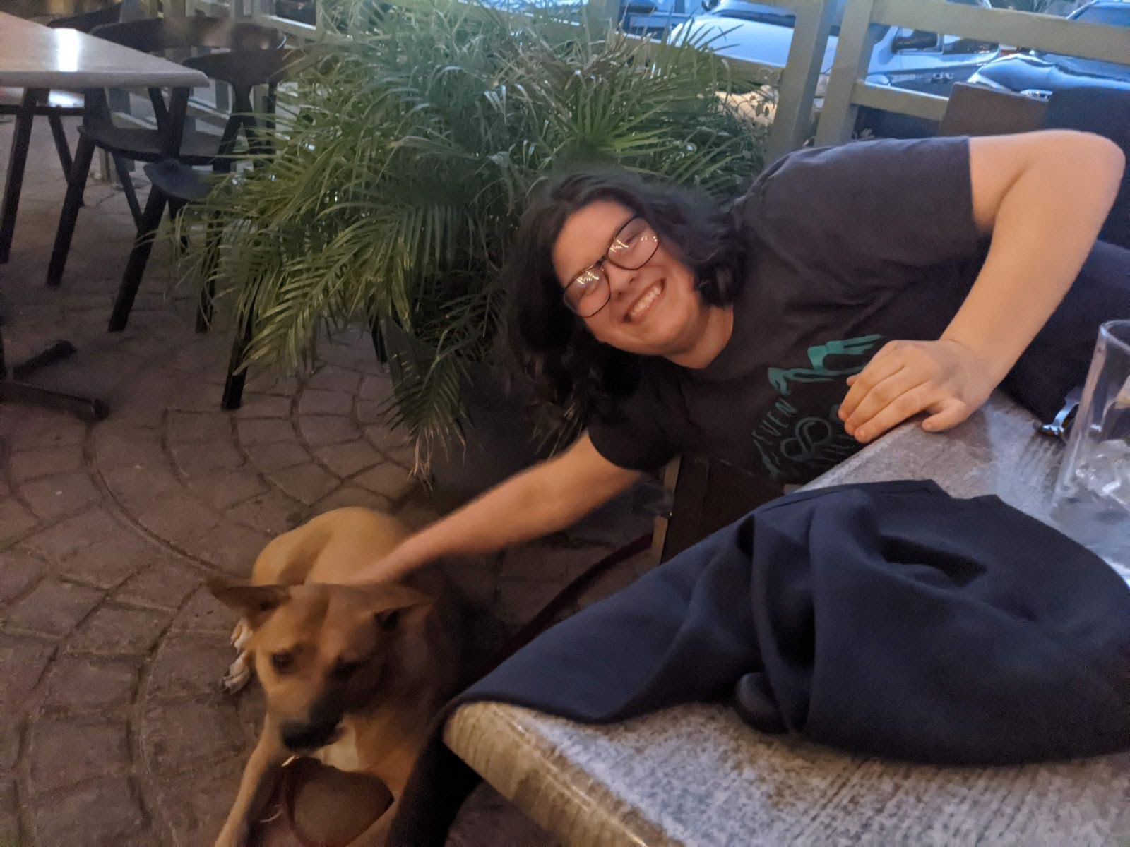 a smiling young man wearing glasses bends over to pet a mediumsized brown dog. they appear to be at a restaurant, as seen by the table, chairs, and presence of a jacket on the table. the lighting is dim and likely from artificial light sources above.