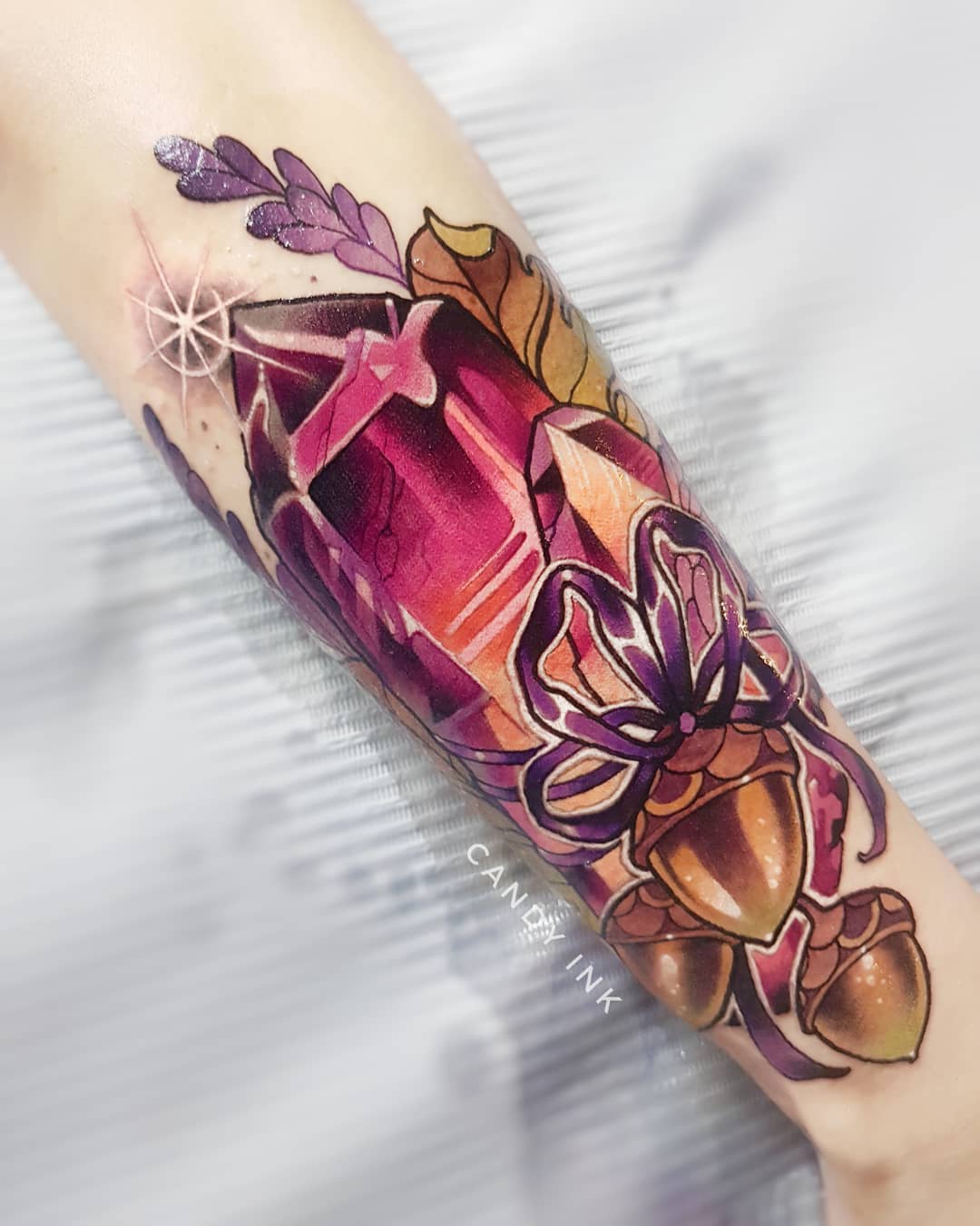 a forearm tattoo on a person's arm. the tattoo is of a purple crystal with a purple bow tied around it. there is a sprig of lavender and two acorns next to the crystal. the tattoo is in the neotraditional style and is very detailed. the colors are vibrant and the overall effect is very striking.