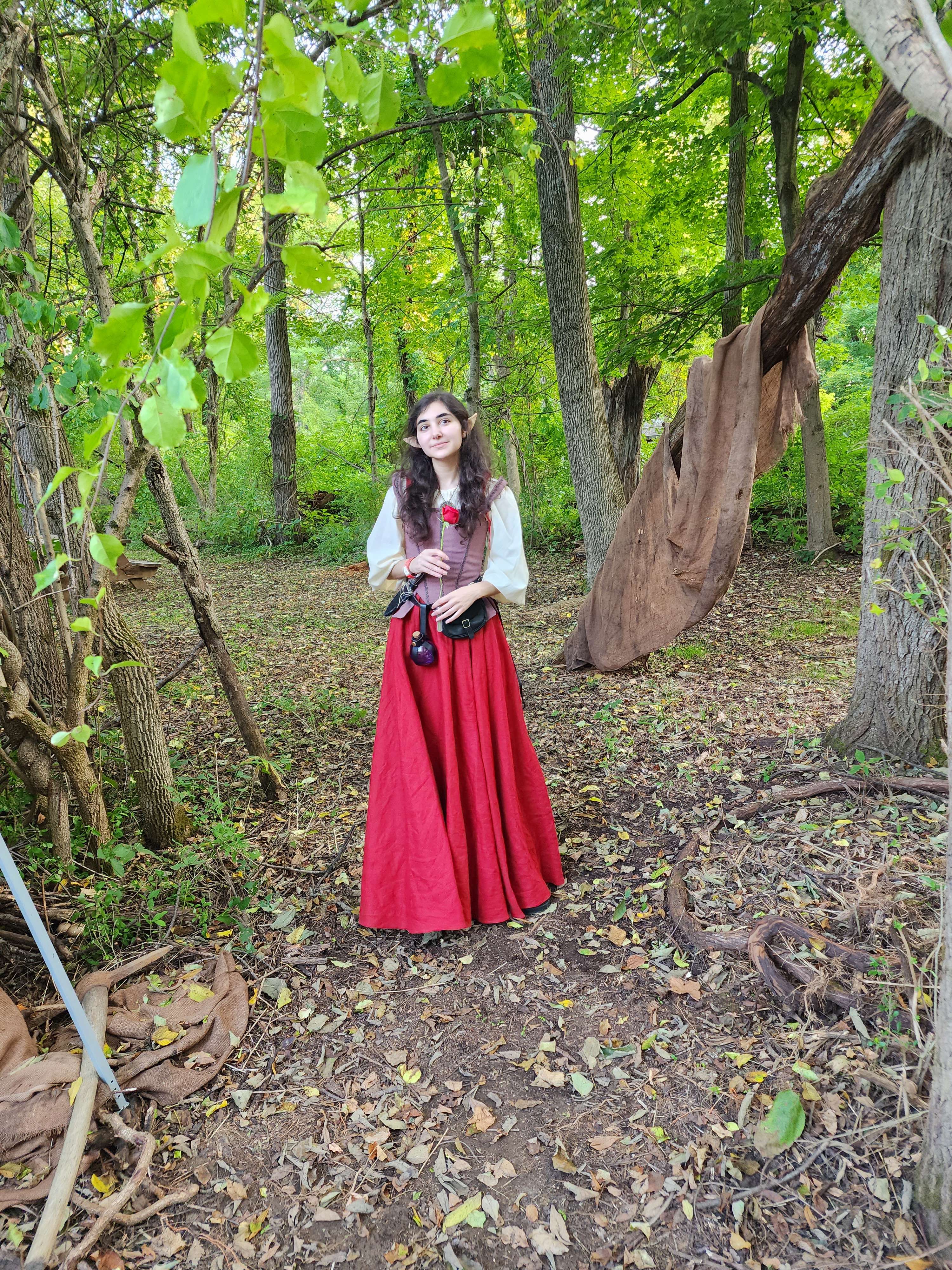 the image shows a young woman dressed in an elf costume. she is wearing a long, flowing red skirt and a light brown, peasantstyle top.  she has pointed ears, indicative of an elf character.  the setting is a forest with many trees and fallen leaves on the ground, suggesting autumn or fall. a large brown fabric is draped over a tree branch. the overall mood is whimsical and fantastical, reminiscent of a renaissance faire or fantasy setting.  she appears to be posing for a photograph.