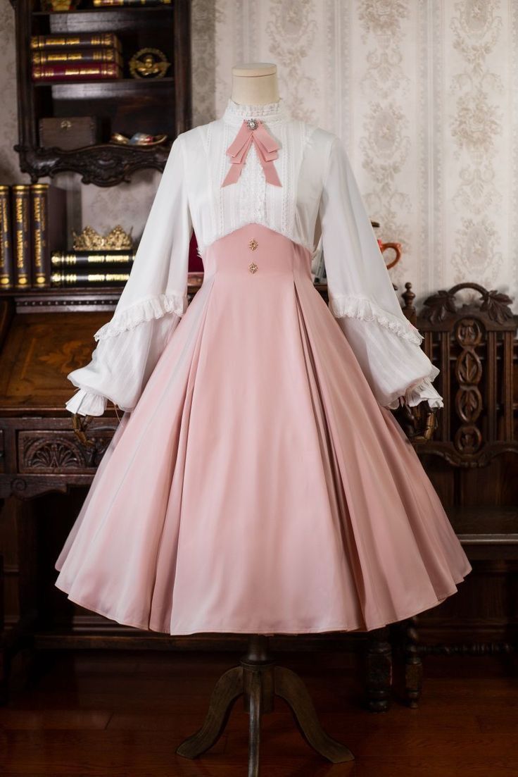 a pink and white lolita fashion dress displayed on a mannequin. the dress features a white blouse with long, flowing sleeves, a pink bow, and a high collar. the pink skirt is full and gathered at the waist. the dress is shown in a room with antique furniture, including a wooden desk and bookshelves. the lighting is bright, illuminating the details of the dress and the room.