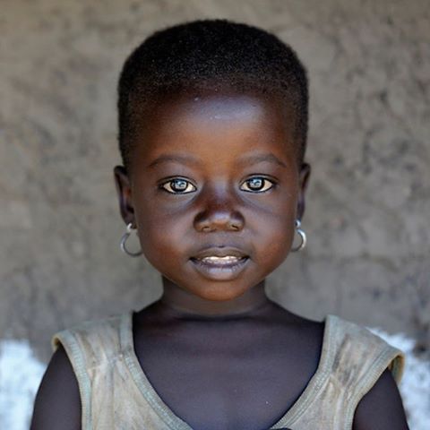 a closeup portrait of a young african girl with dark skin, short hair, and silver hoop earrings. she is wearing a tan top and is standing in front of a textured gray wall.