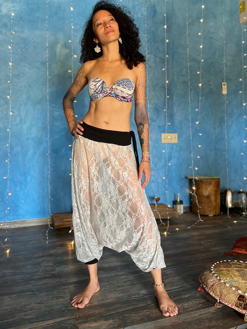 a fullbody shot of a woman with long curly dark hair and tattoos posing in a light creamcolored lace harem pants outfit. she is barefoot and wearing a black waistband over a matching top.  her top is a strapless floral bra. the backdrop is a blue wall decorated with string lights, and the setting is an indoor space with a wooden floor and some floor pillows visible in the bottom right. the overall style is bohemian and suggests a potential connection to dancewear or yoga clothing.