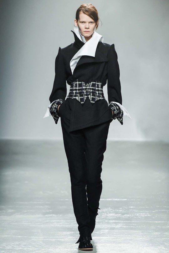 a woman in a black jacket with a white collar and a plaid belt walks down a runway. she is wearing black pants and black boots.