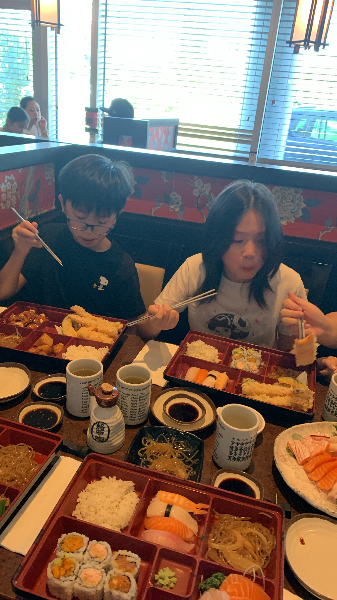 he image shows two children enjoying a japanese meal at a restaurant. the children are seated at a table with several bento boxes filled with various japanese dishes, including sushi, tempura, rice, and vegetables. the scene is welllit with natural light coming through the window behind them. the overall atmosphere is warm and inviting.
