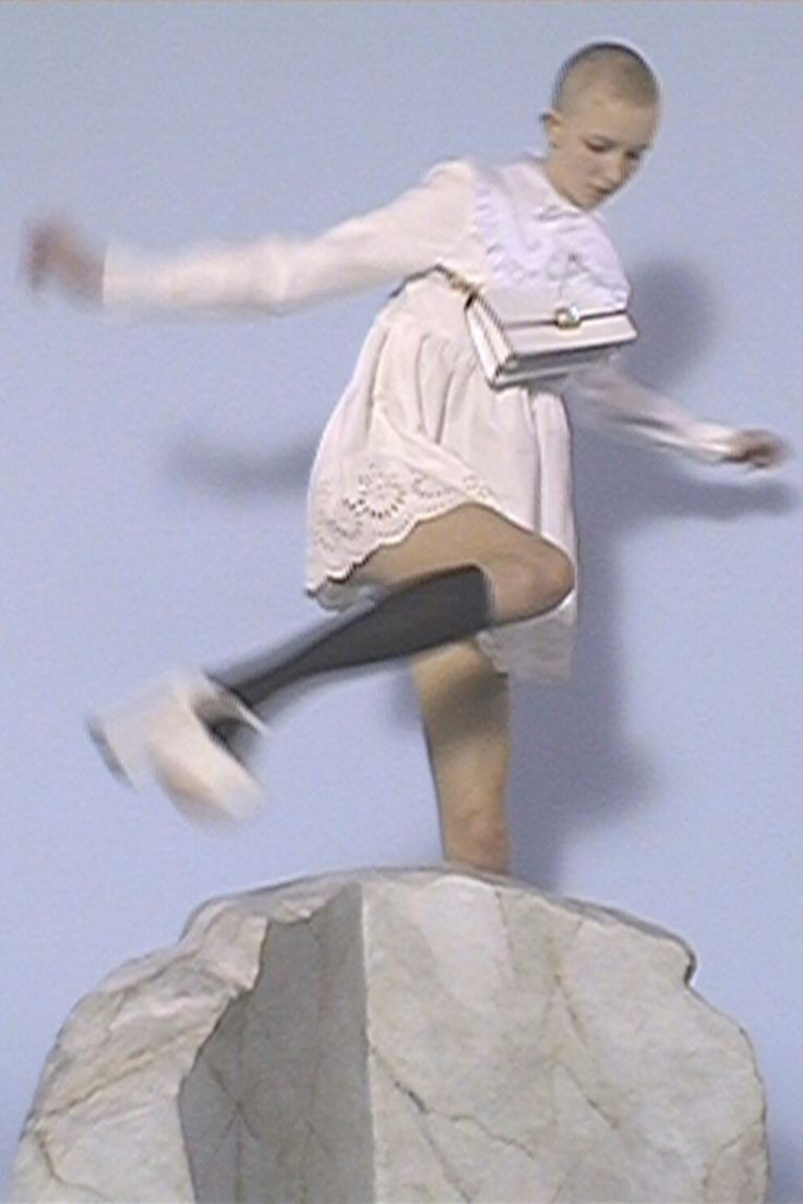 a young woman wearing a white dress and black boots poses on a large rock while holding a small white purse.